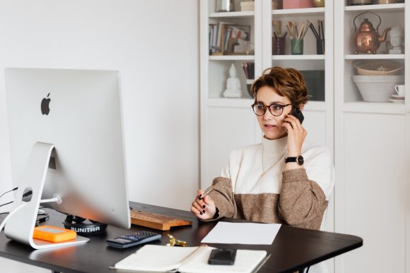 Kvinna i glasögon sitter vid skrivbord och pratar i telefon